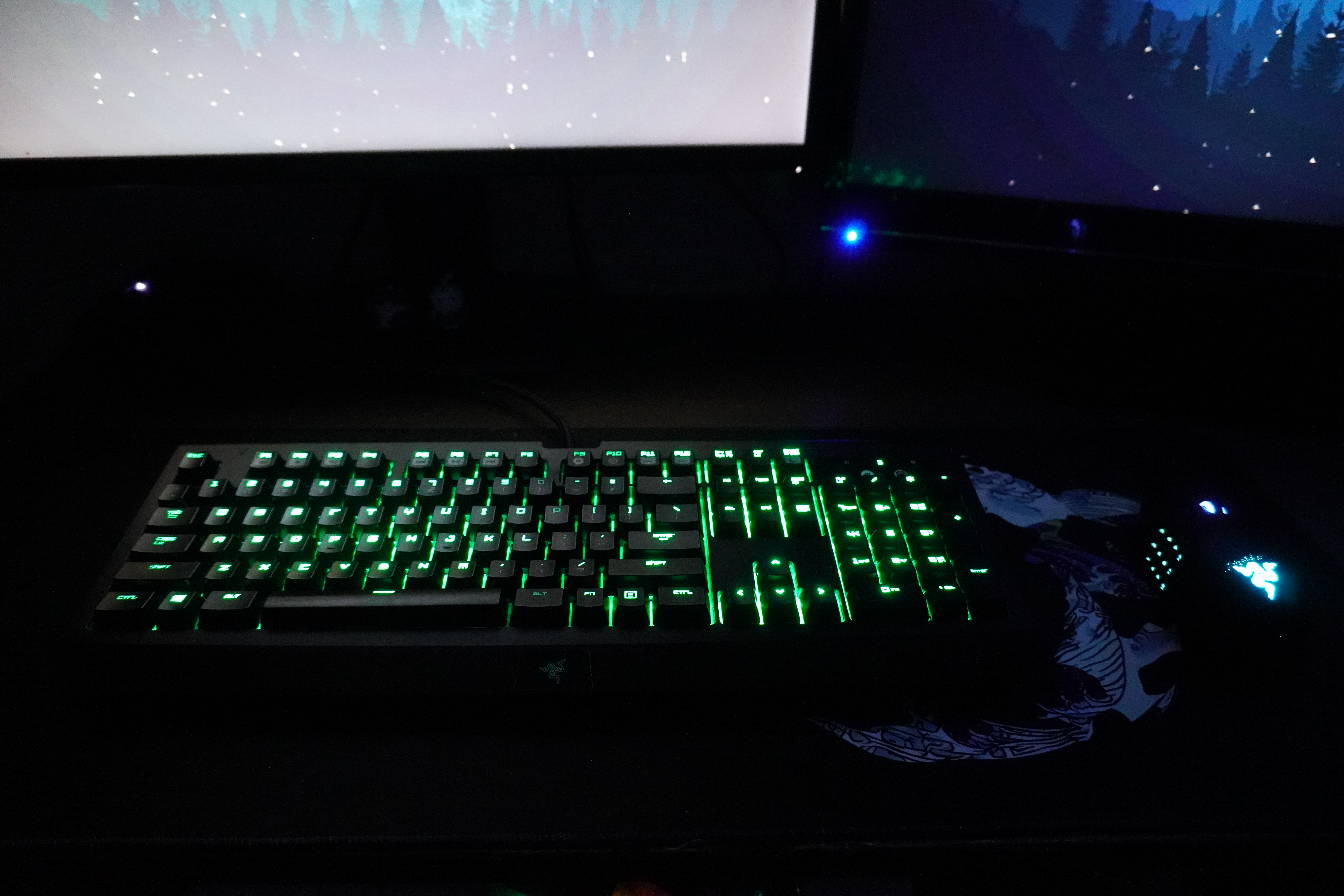 Razer BlackWidow Ultimate 2016 with green backlighting beneath two monitors, with a mouse on a patterned mousepad to the right.